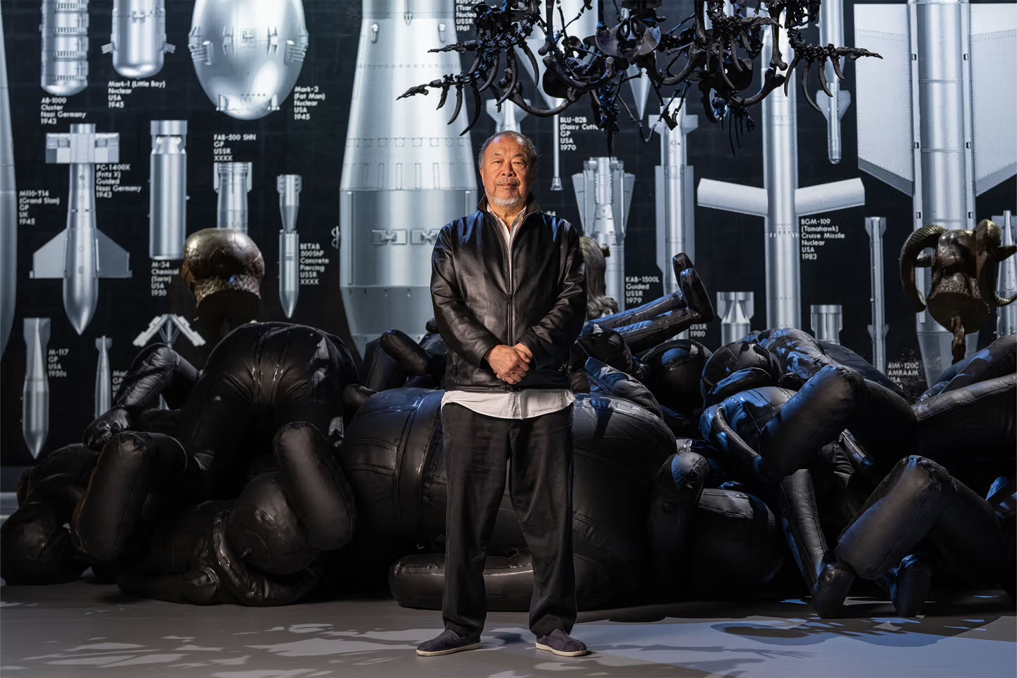 Photographie diurne de studio en plan pied de Ai Weiwei debout au centre de la scène de l'installation Button Up aux Aviva Studios de Manchester en juillet 2026, artiste chinois vêtu d'une veste en cuir noir sur chemise blanche et pantalon noir cheveux et barbe grisonnants regard tourné vers l'objectif, disposé devant un amoncellement au sol de silhouettes gonflables noires en vinyle représentant des corps recroquevillés, immense chandelier baroque suspendu au plafond composé de squelettes noirs et branches osseuses entrelacées, arrière-plan mural en noir imprimé d'une planche technique blanche d'inventaire de missiles armes atomiques et bombes conventionnelles avec inscriptions Mark-1 Little Boy USA 1945 Mark-2 Fat Man Fritz X Guided Nazi Germany BLU-82B Daisy Cutter USA 1970 BGM-109 Tomahawk USA 1983, sol clair balayé de reflets, éclairage direct frontal spot dur