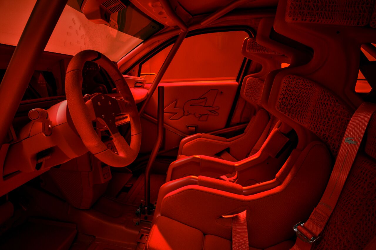 Vue intérieure côté conducteur de la A290 Rallye Alpine Lacoste « Beware of the crocodile », habitacle entièrement rouge écarlate baigné d'une lumière rasante, volant sport à trois branches, quatre sièges baquets en textile petit piqué Lacoste rouge, harnais quatre points rouges, arceau de sécurité intégral, portière ornée d'un large crocodile embossé et du A d'Alpine, mailles techniques 3D visibles sur l'arrière des sièges, ambiance de tapisserie technique où chaque surface est habillée façon polo haute couture