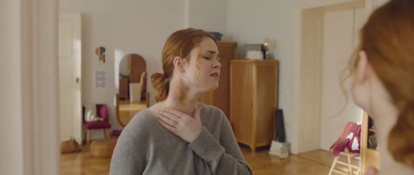 Photogramme du film Three Words signé Publicis Conseil pour AXA récompensé par le Titanium Grand Prix des Cannes Lions 2026, plan rapproché d'une jeune femme rousse en pull gris debout dans un appartement parisien, main droite portée vers la gorge où apparaissent des marques rouges de strangulation, regard détourné vers la droite, intérieur soigné avec armoire en bois et fauteuil rose framboise en arrière-plan, lumière naturelle douce