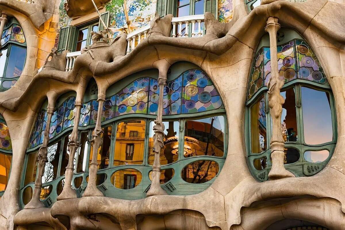Photographie en contre-plongée serrée sur la façade principale de la Casa Batlló d'Antoni Gaudí à Barcelone chef-d'œuvre du modernisme catalan qui a inspiré la typographie du FC Barcelone Modernista, gros plan sur les fenêtres organiques du bel étage aux montants en pierre sculptée en forme d'os aux courbes fluides et végétales entourant des ouvertures en arc surbaissé et des vitraux en disques polychromes bleus verts violets et jaunes, garde-corps en fer forgé courbé et balcons cylindriques en pierre grise, façade dorée réfléchie dans les vitres et détail de mosaïque de trencadís aux tons verts et jaunes en haut de cadre, scène urbaine complètement différente de la photo produit du maillot