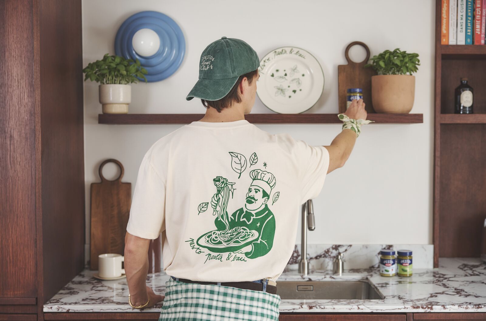 Photographie d'ambiance du Basil Bar Barilla dévoilée à l'occasion du pop-up Étienne-Marcel du 19 au 28 juin 2026, jeune homme de dos dans une cuisine ouverte portant un t-shirt blanc cassé décoré au feutre vert d'une illustration de chef italien soulevant une assiette de pâtes signée Pesto, Pasta and Love, casquette en velours côtelé vert basilic vissée sur la tête, foulard vert glissé à la ceinture, sur la pointe des pieds pour attraper un pot Pesto Genovese Barilla rangé sur une étagère en bois sombre garnie de plantes aromatiques