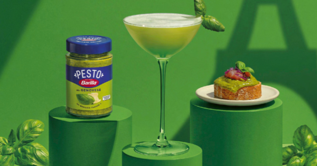 Composition produit lumineuse du Basil Bar Barilla sur fond uni vert basilic, podium en deux niveaux peints du même vert que l'arrière-plan, à gauche un pot de Pesto Genovese Barilla à étiquette bleu marine et liseré bleu posé sur une marche basse, au centre un verre à martini empli d'un cocktail vert basilic mousseux décoré d'une feuille de basilic frais accrochée à la coupe, à droite sur le second podium une assiette ronde blanche supportant une bruschetta dorée garnie de pesto épais et de tomates confites, feuilles de basilic vert vif disposées au premier plan en bas du cadre