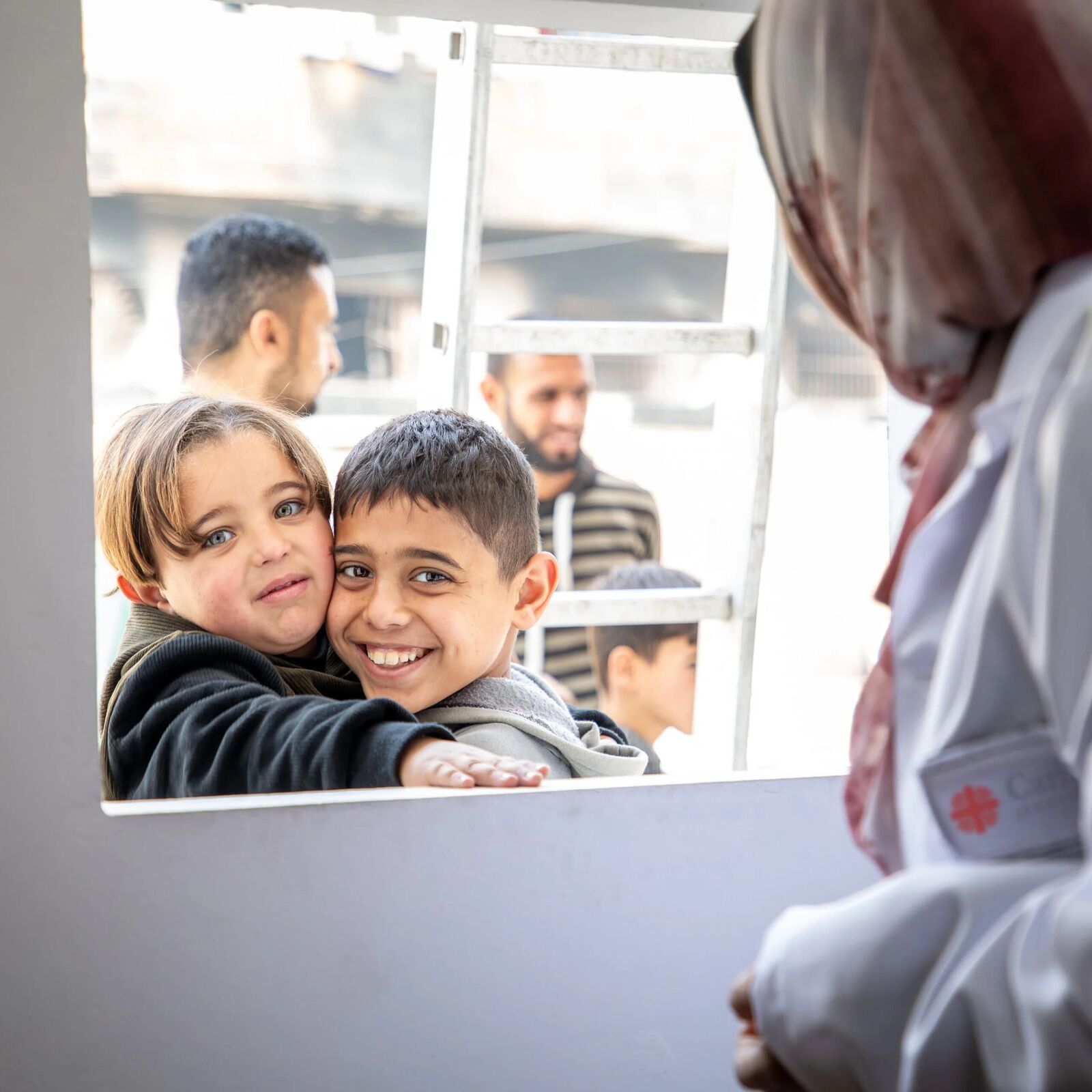 Image dévoilée par Caritas Jérusalem en mai 2026 de l'intérieur du dispensaire Vehicle of Hope, deux enfants vus depuis l'intérieur du véhicule par la baie vitrée du compartiment de soin, un petit garçon brun en pull noir et une petite fille en pull gris-marron se serrent en souriant devant la fenêtre, paroi blanche du compartiment médical avec discret logo Caritas brodé en rouge sur l'épaule droite d'une soignante en blouse blanche et hijab beige au premier plan, à l'arrière-plan d'autres enfants et adultes attendent leur tour dans une ruelle bétonnée de Gaza