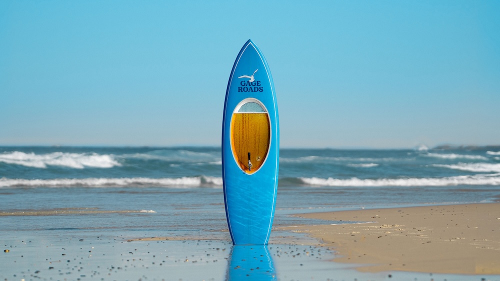 Visuel principal de la campagne Beerboard dévoilée par la brasserie indépendante australienne Gage Roads Brew Co le 30 juin 2026 pour sa Summer Ale Single Fin, planche de surf bleu ciel dressée à la verticale sur le sable mouillé d'une plage de Perth au bord de l'océan Indien, chambre transparente ovale au centre de la board révélant les 10 litres de bière ambrée avec un robinet cuivré, mention Gage Roads sérigraphiée en noir au sommet sous une mouette au trait, vagues blanches en toile de fond et ciel bleu limpide