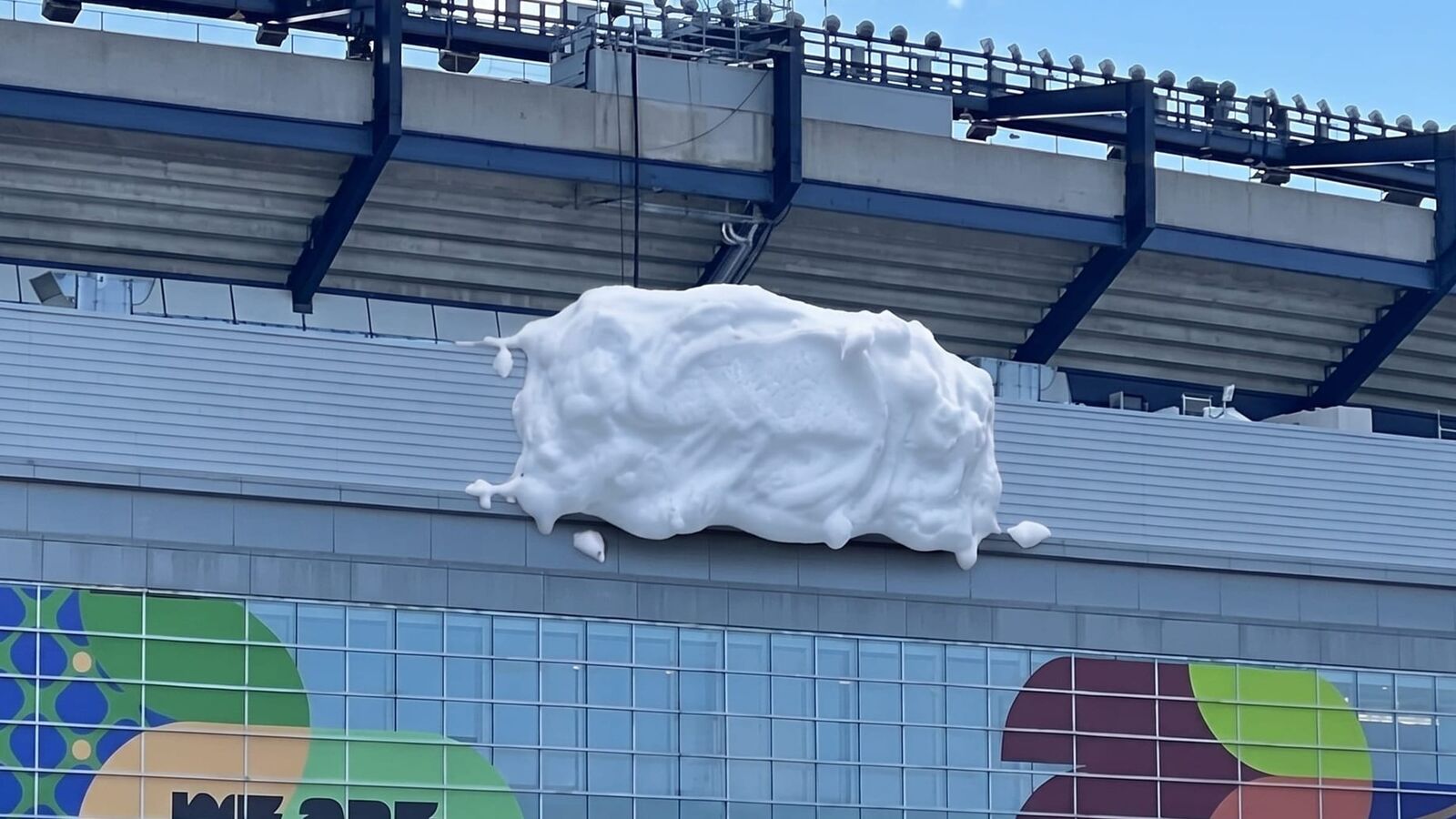 Vue aérienne du Gillette Stadium dévoilée le 21 juin 2026 pendant la Coupe du Monde 2026, terrain de football vert vif vu du ciel entouré de 64 146 sièges chacun recouvert d'un sticker bleu imprimé en faux relief de mousse à raser blanche dégoulinante