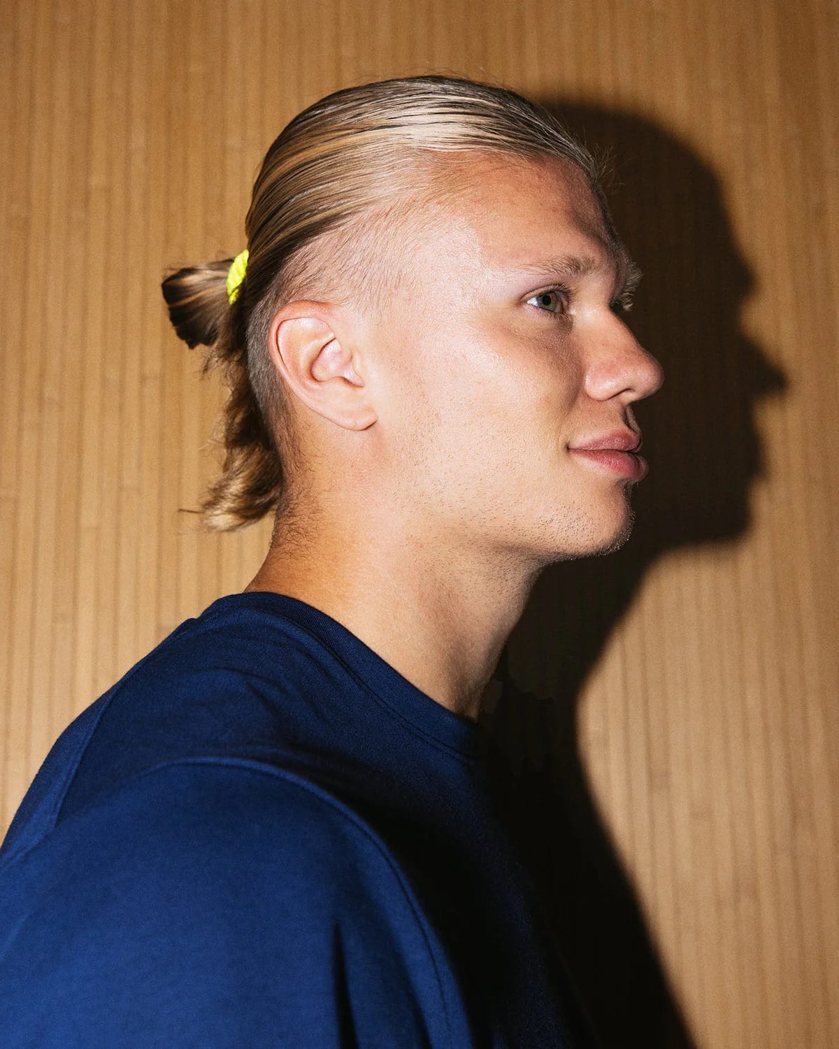 Photographie éditoriale extraite de la campagne d'affichage Haaland Edition de Kknekki à Oslo à partir du 29 juin 2026, portrait en profil serré du footballeur Erling Haaland cheveux blonds plaqués en arrière et coiffés en un chignon bas tenu par un élastique Kknekki jaune fluo, mâchoire nette et regard tourné vers la droite, tee-shirt bleu marine, arrière-plan en panneaux de bois clair légèrement striés, ombre portée nette contre le mur suggérant un éclairage latéral chaud
