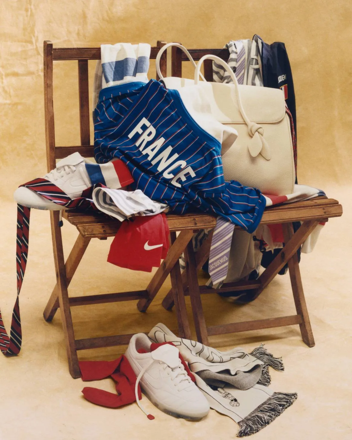 Nature morte officielle de la collection Jacquemus x Nike x FFF posée sur une chaise en bois, maillot bleu FRANCE plié, sac tote, sneakers blanches, écharpe et accessoires aux couleurs des Bleus, photographiée pour le lancement du 11 juin 2026 et diffusée par Fashion United