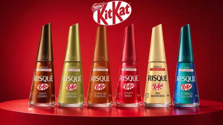 Photo de campagne officielle de la collection capsule Risqué x KitKat dévoilée au Brésil le 30 juin 2026, plan produit en studio sur fond rouge dégradé avec logo KitKat sérigraphié en blanc et rouge sur bandeau supérieur, alignement horizontal parfaitement calibré de six flacons de vernis à ongles Risqué de 8 ml aux bouchons pyramidaux colorés dans l'ordre brun métallique Choco Cósmico, or crémeux Crocante na Medida, cuivre orangé Só Mais Um Pedacinho, rouge métallique KitKat Mania, beige nude Meu Wafervorito et vert émeraude à effet Eu Mereço Esse Glow, étiquette Risqué blanche et bandeau Metálico Com Cheirinho sur chaque flacon, socle vermillon lumière rasante