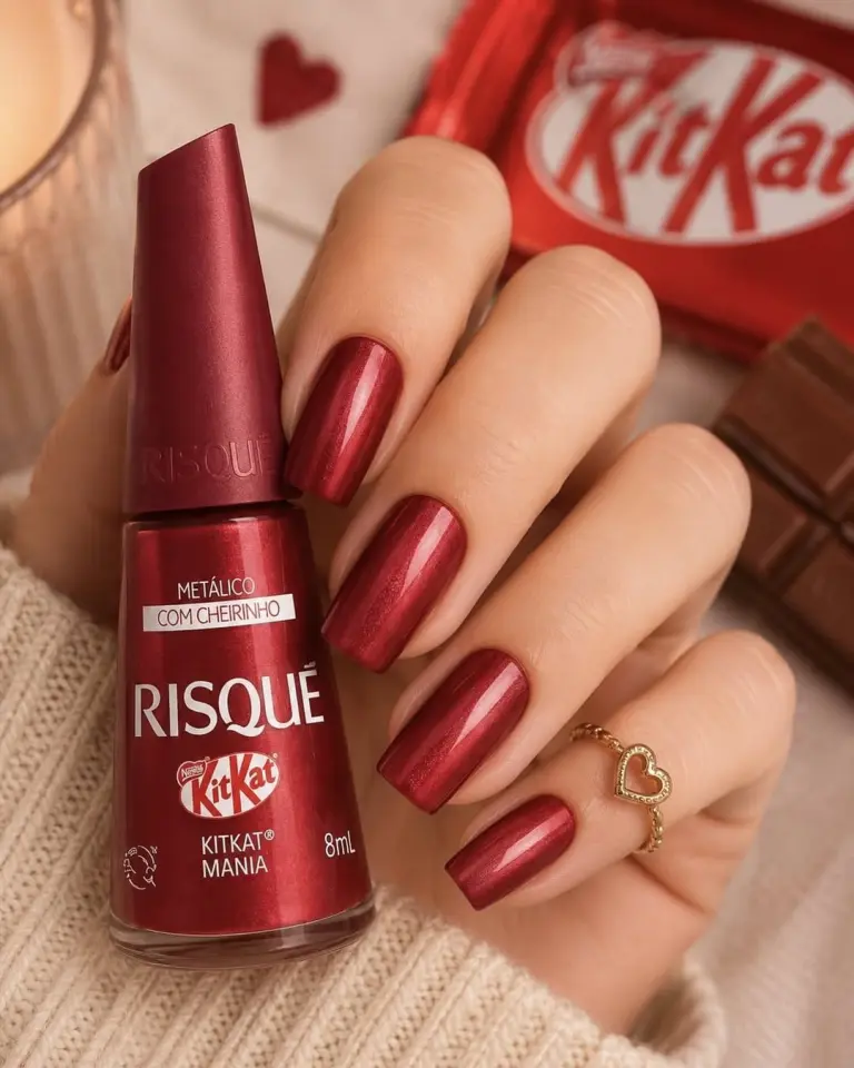 Photographie lifestyle rapprochée diffusée fin juin 2026 pour la collection Risqué x KitKat, main de femme aux ongles longs carrés soigneusement vernis en rouge métallique brillant KitKat Mania avec bague fine en cœur doré tenant à la verticale un flacon Risqué de 8 ml au capot rouge burgundy portant l'étiquette KitKat Mania Metálico Com Cheirinho posé contre un pull tricoté ivoire, en arrière-plan flou une bougie beige allumée et un emballage KitKat rouge original froissé aux côtés d'un morceau de tablette au chocolat noir posé sur un bristol blanc décoré de deux cœurs rouges