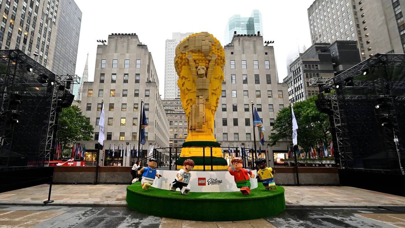 Photographie en plan large diurne à hauteur de rue au cœur de Rockefeller Plaza à New York le 7 juillet 2026, immense réplique du trophée de la Coupe du Monde FIFA entièrement construite en briques LEGO jaunes et vertes s'élevant à 8 mètres avec son socle de gazon synthétique posé sur une estrade blanche sérigraphiée LEGO Editions, quatre minifigurines LEGO grandeur nature de joueurs de football floqués 10 et 7 en tenues bleue argentine portugaise rouge et brésilienne jaune plantées autour du socle, drapeaux des pays participants alignés sur mâts blancs autour de l'installation, immeubles Art déco pierre claire du Rockefeller Center encadrant la scène en fond, ciel gris pommelé de fin d'après-midi et projecteurs sur portiques noirs latéraux, sol pavé mouillé