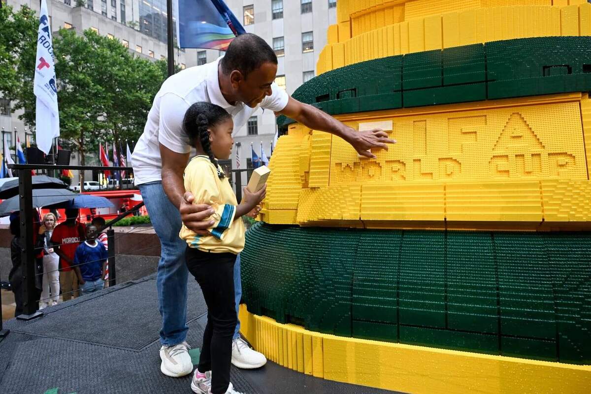 Photographie en gros plan diurne au pied de la sculpture LEGO du trophée de la Coupe du Monde installée à Rockefeller Plaza à New York le 7 juillet 2026, la légende brésilienne Cafu chemise polo blanche jean bleu cheveux courts châtain penché à gauche présente à une petite fille en maillot jaune du Brésil aux cheveux tressés en couette basse une brique jaune posée sur le mot FIFA en construction dans le socle vert et jaune de la sculpture géante montrant les lettres WORLD CUP sculptées en briques, autour deux enfants en maillots rouge et bleu observent la scène derrière une balustrade en câbles noirs, drapeaux nationaux flottants et immeubles art déco en arrière-plan flou, image de dévoilement officiel de la LEGO Fan Zone