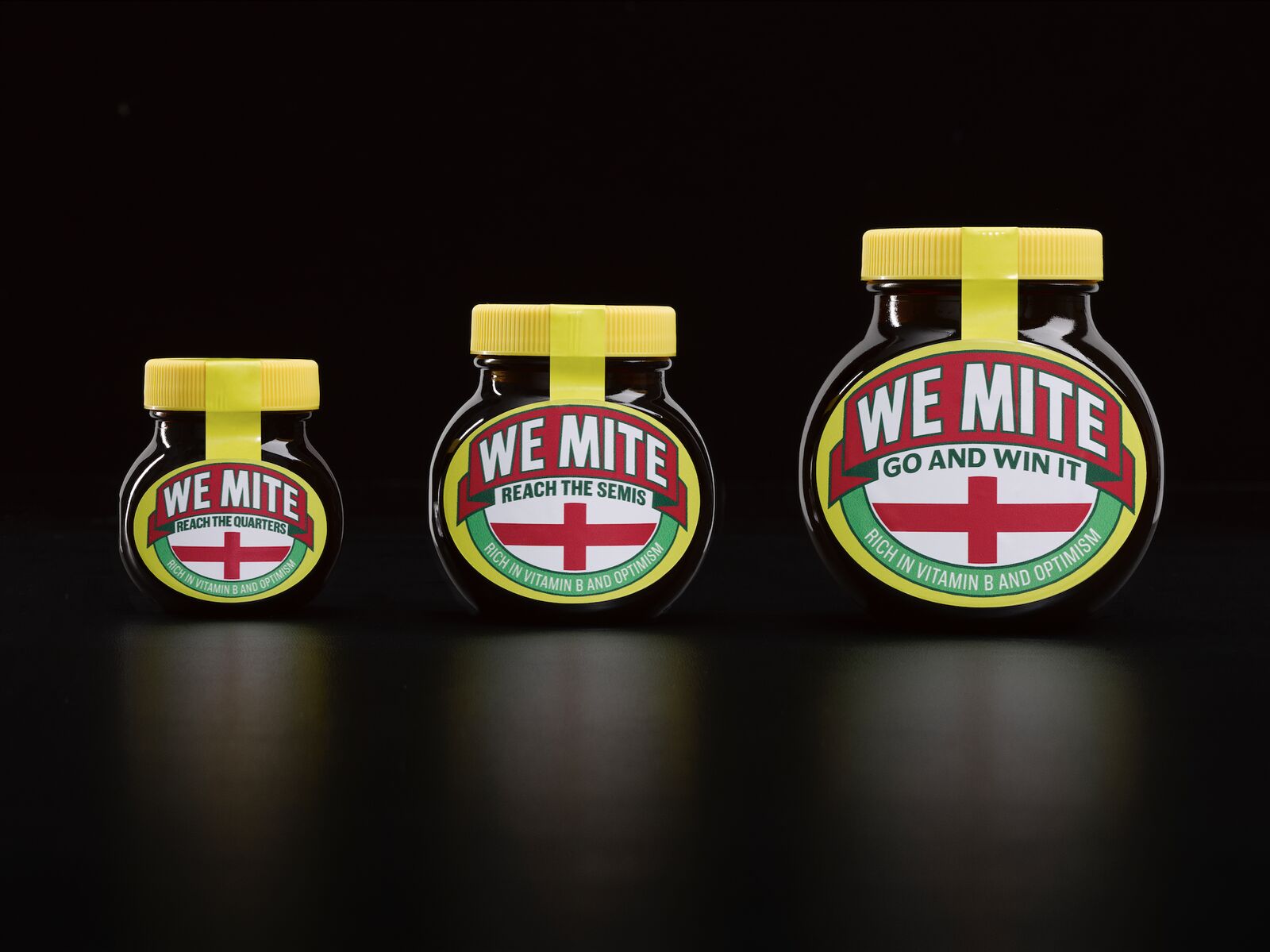 Packshot studio officiel des trois pots WeMite en édition limitée lancés par Marmite le 1er juillet 2026 pour la Coupe du monde de football, cadrage panoramique sur fond noir profond, à gauche petit pot 125 g étiquette ovale rouge et verte estampillée WE MITE REACH THE QUARTERS avec drapeau anglais croix rouge sur fond blanc et mention Rich in vitamin B and optimism, au centre pot moyen 250 g même étiquette avec la mention REACH THE SEMIS, à droite grand pot 500 g avec la mention GO AND WIN IT, tous coiffés du bouchon jaune vif signature et de la bande de scellage jaune, ombres portées réfléchies sur la surface laquée sombre