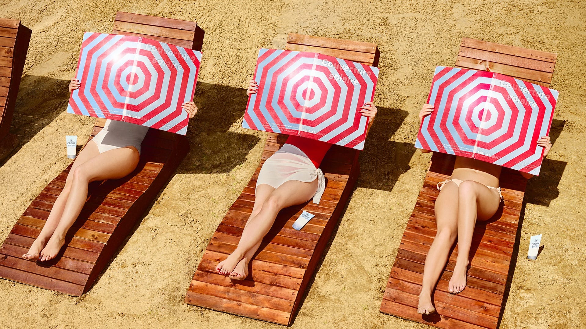 Photographie de plage diurne en vue de dessus zénithale de la campagne Couverture Solaire de Melanoma Canada signée Rethink en juillet 2026, cadrage horizontal centré sur trois transats en bois foncé alignés côte à côte disposés en oblique sur le sable clair d'une plage, sur chaque transat une femme allongée cachant son visage et son buste derrière une grande couverture de livre rectangulaire géante ouverte au motif de parasol rouge et blanc en cercles concentriques éblouissant reflet du soleil et petits textes en pied, femmes en maillots deux-pièces beige rouge et beige avec sarongs blanc et beige, un flacon blanc de crème solaire SPF 50 posé au sol à côté de chaque transat, ombres portées nettes des transats sur le sable, éclairage vertical solaire dur de midi