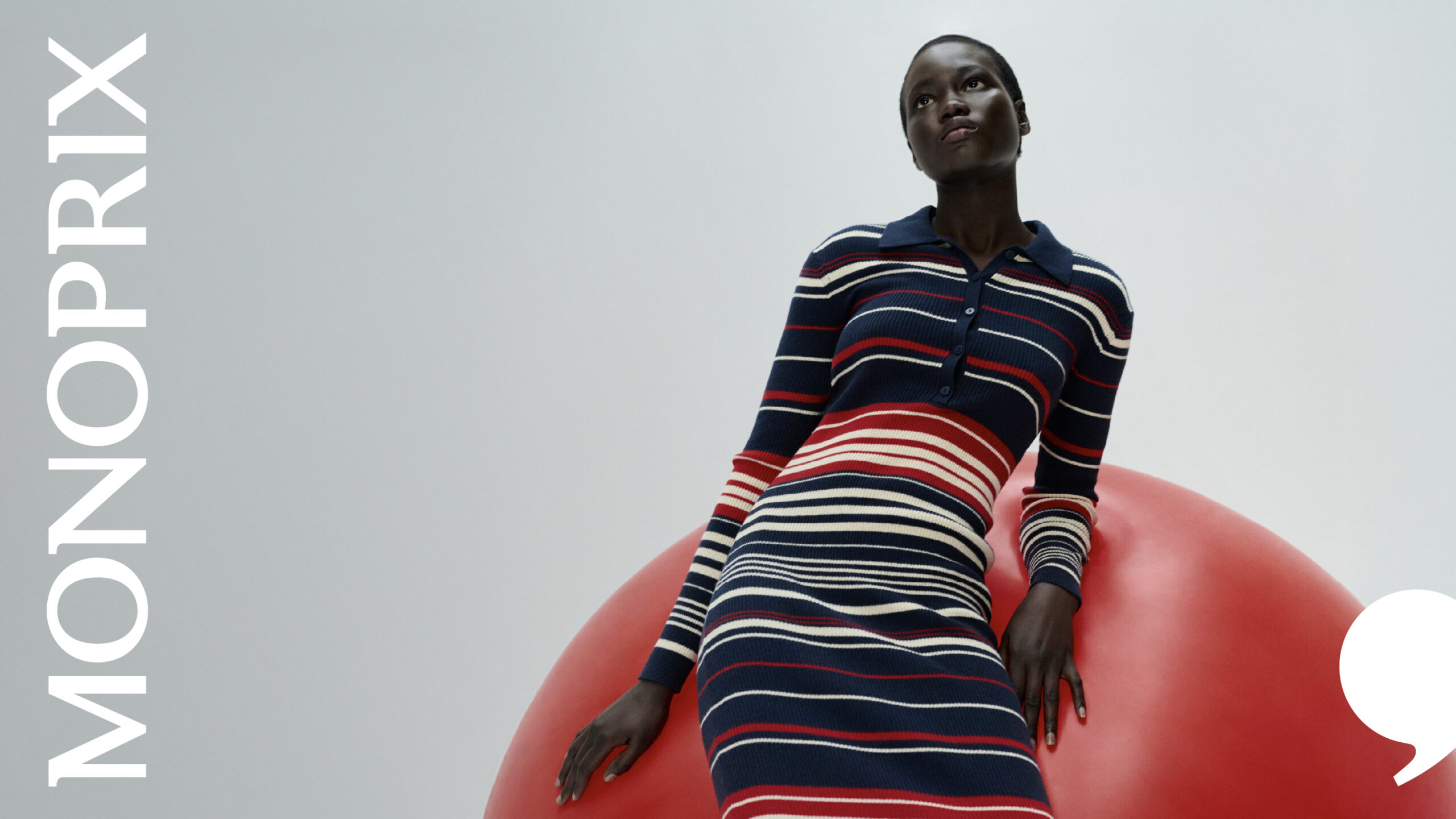 Visuel campagne de la nouvelle identité Monoprix signée W Conran Design, photographie de studio à cadrage horizontal élégant sur fond gris blanc dégradé neutre, mannequin femme à peau noire cheveux courts noir aile de corbeau en robe polo long boutonnée à rayures horizontales larges bleu marine crème et rouge écarlate, corps assis en équilibre sur une grande boule gonflable rouge coquelicot ronde et brillante posée à droite du cadre, mains posées sur la sphère bras tendus regard concentré porté vers le haut à gauche, mot MONOPRIX en très grands caractères empattés blancs disposé verticalement le long du bord gauche du cadre en composition typographique éditoriale, virgule signature Monoprix en blanc placée en bas à droite du cadre, ambiance esthétique magazine mode française des années 1970 revisitée pop et contemporaine