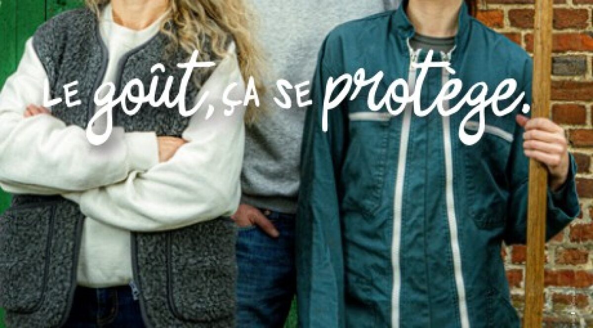 Visuel principal de la campagne d'affichage Le goût ça se protège signée Asgard pour les Fromages AOP de Normandie dévoilée le 17 juin 2026, deux portraits cadrés au buste d'éleveurs normands côte à côte devant un mur de briques rouges et un volet vert, l'une porte un gilet en laine grise sur un pull beige et croise les bras, l'autre porte une veste de travail vert sapin et tient une fourche en bois, signature manuscrite blanche LE GOÛT ÇA SE PROTÈGE incrustée en surimpression sur les vêtements