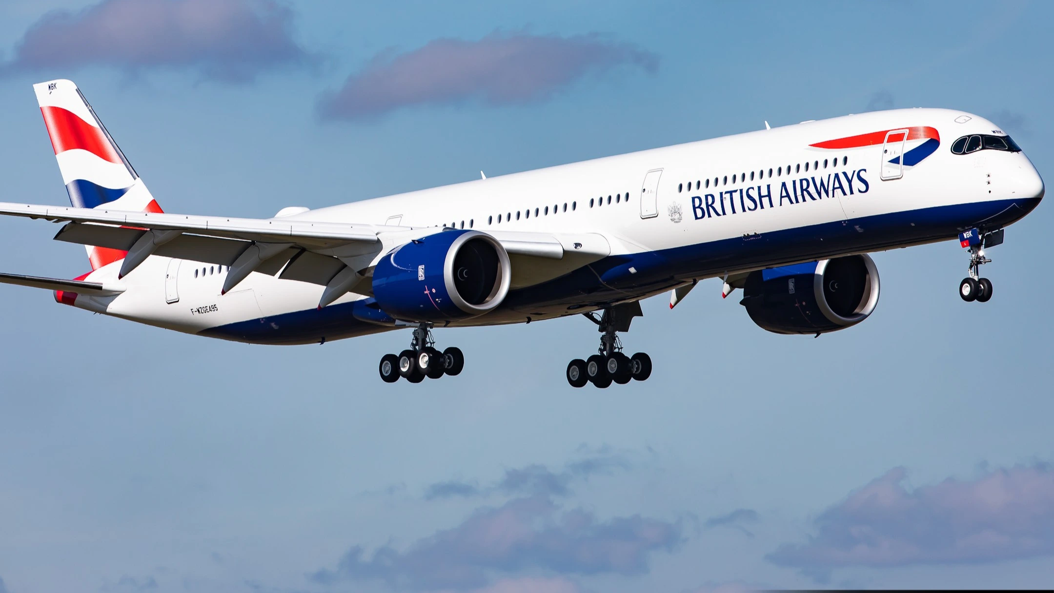 Photographie de spotting aviation en plongée moyenne diurne de l'Airbus A350 1041 immatriculé F-WZGE de British Airways en phase d'approche finale, long fuselage blanc éclatant avec inscription bleu marine BRITISH AIRWAYS en gros caractères sur la partie centrale droite, cockpit et nez au drapeau britannique Union Jack stylisé aux couleurs rouge bleu et blanc balayant le nez et se prolongeant en vagues sur la queue verticale de l'appareil, deux gros moteurs Rolls-Royce Trent XWB gris satiné sous les ailes en position basse, train d'atterrissage sorti avec roulettes noires alignées, ciel bleu pâle strié de nuages bas roses cotonneux au coucher du soleil, atmosphère cinématographique d'aéroport international