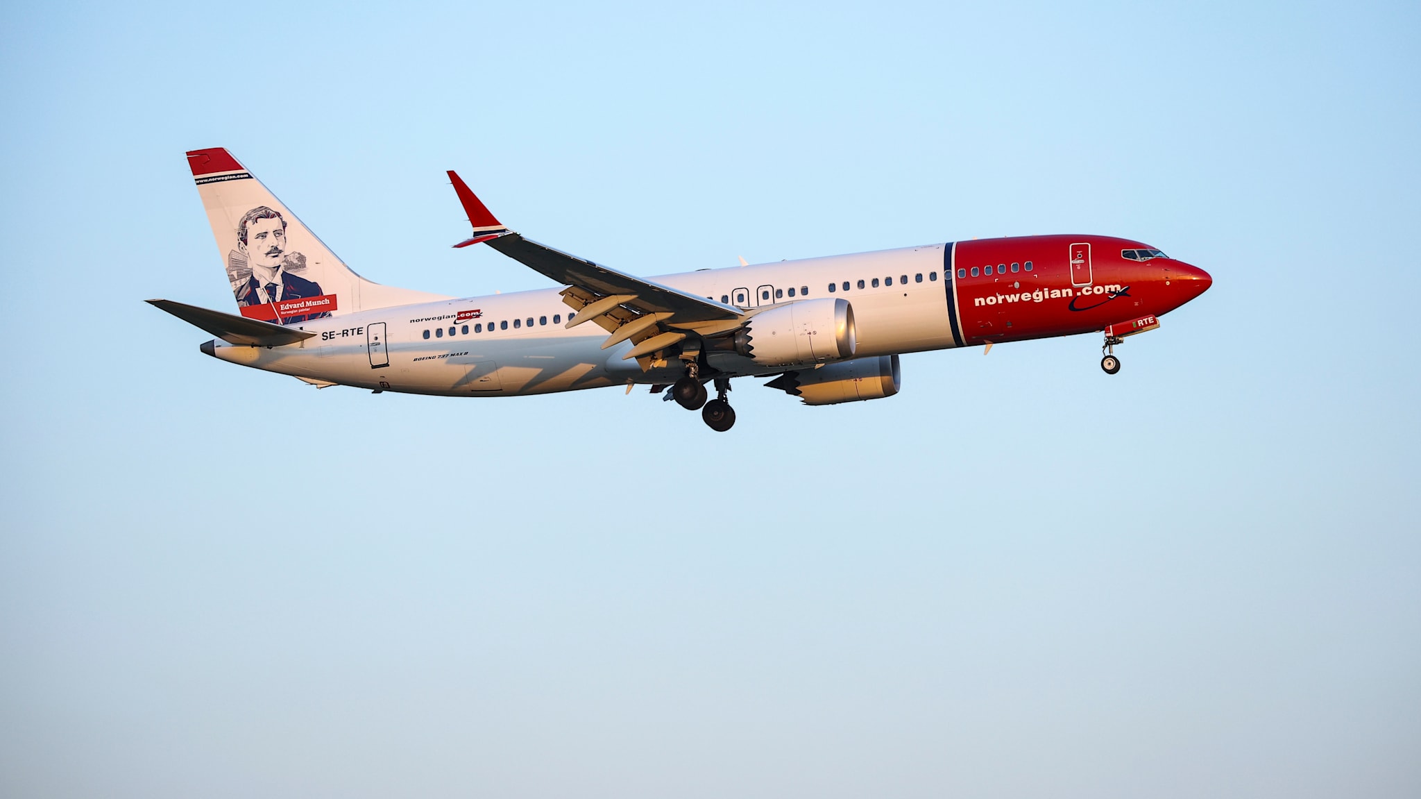 Photographie de spotting aviation en léger contre-plongée diurne d'un Boeing 737 MAX 8 immatriculé SE-RTE de la compagnie Norwegian Air Shuttle en phase d'atterrissage, fuselage blanc satiné aux hublots alignés avec inscription rouge norwegian.com sur le nez de l'appareil et grand rouge écarlate de la partie avant du fuselage, dérive verticale blanche portant un portrait dessiné en noir et blanc du peintre norvégien Edvard Munch surmonté d'un liseré rouge et bleu au motif du drapeau norvégien, moteur CFM Leap noir et gris clair sous l'aile droite, ciel bleu ciel dégradé de lever de soleil, sans nuage, éclairage doré chaud de fin de journée