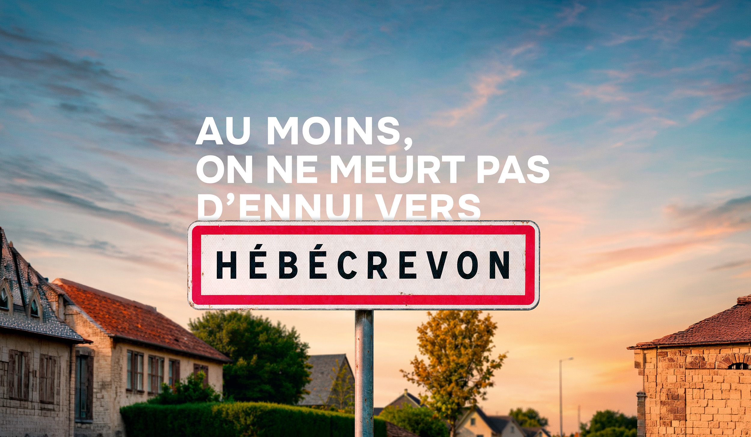 Affiche verticale JCDecaux de la nouvelle campagne PagesJaunes signée Marcel déployée à partir du 10 juillet 2026, panneau routier d'entrée d'agglomération encadré de rouge sur fond blanc portant le nom de la commune HÉBÉCREVON planté au premier plan devant un ciel de fin de journée aux nuances roses et bleues, en arrière-plan un village normand avec toits de tuiles rouges maison en pierre calcaire claire et façades chaulées, accroche AU MOINS ON NE MEURT PAS D'ENNUI VERS HÉBÉCREVON en grandes capitales blanches en haut du visuel, format print vertical 120x176