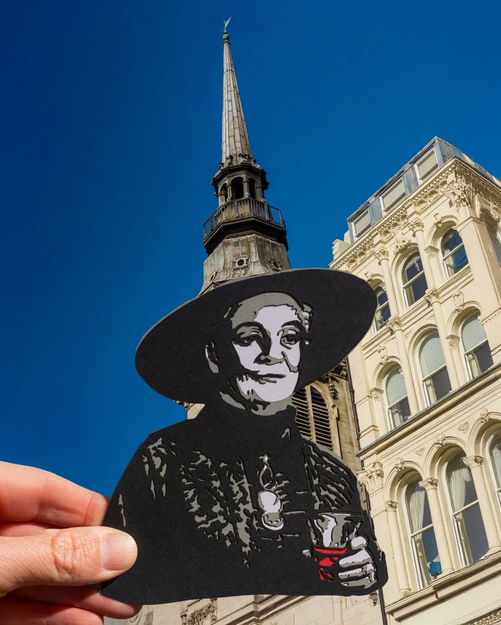 Deuxième photographie Paperboyo mise en avant le 19 juin 2026, plan rapproché frontal de Big Ben à Londres ciel bleu pâle d'hiver, une main au premier plan tient une découpe en papier noir représentant la silhouette d'un gobelet à milkshake avec sa paille qui s'aligne parfaitement avec la tour horloge laissant croire qu'on aspire le sommet de Big Ben comme une boisson, photographie en lumière naturelle de matin sans aucune retouche