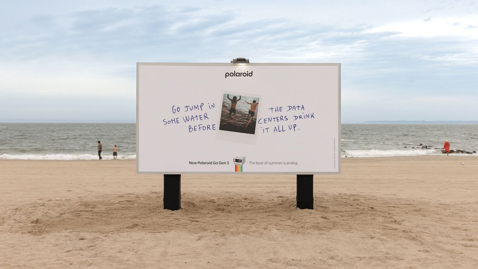 Panneau publicitaire géant Polaroid The Best of Summer Is Analog dévoilé le 19 juin 2026 planté directement dans le sable doré de la plage de Coney Island à New York, format horizontal blanc cassé avec le slogan en lettres capitales noires Go jump in some water before the data centers drink it all up, logo arc en ciel Polaroid en bas, ciel bleu pâle d'été, parasols rayés multicolores et baigneurs en arrière-plan flou