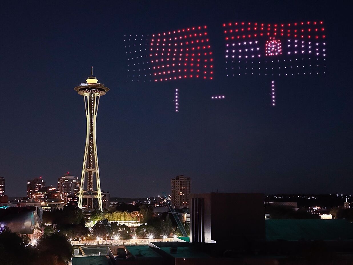 Photo de nuit du premier scoreboard aérien mondial signé Visit Seattle diffusé le 19 juin 2026 au-dessus de Seattle Center pour la rencontre États-Unis Australie de la Coupe du monde FIFA 26, cadrage large sur la Space Needle jaune lumineuse au premier plan à gauche et sur la ligne d'horizon de Seattle en contrebas, dans le ciel bleu nuit 400 drones lumineux forment simultanément le drapeau américain à gauche et le drapeau australien à droite avec l'inscription pointillée du score de la rencontre en petits chiffres blancs suspendus entre les deux drapeaux