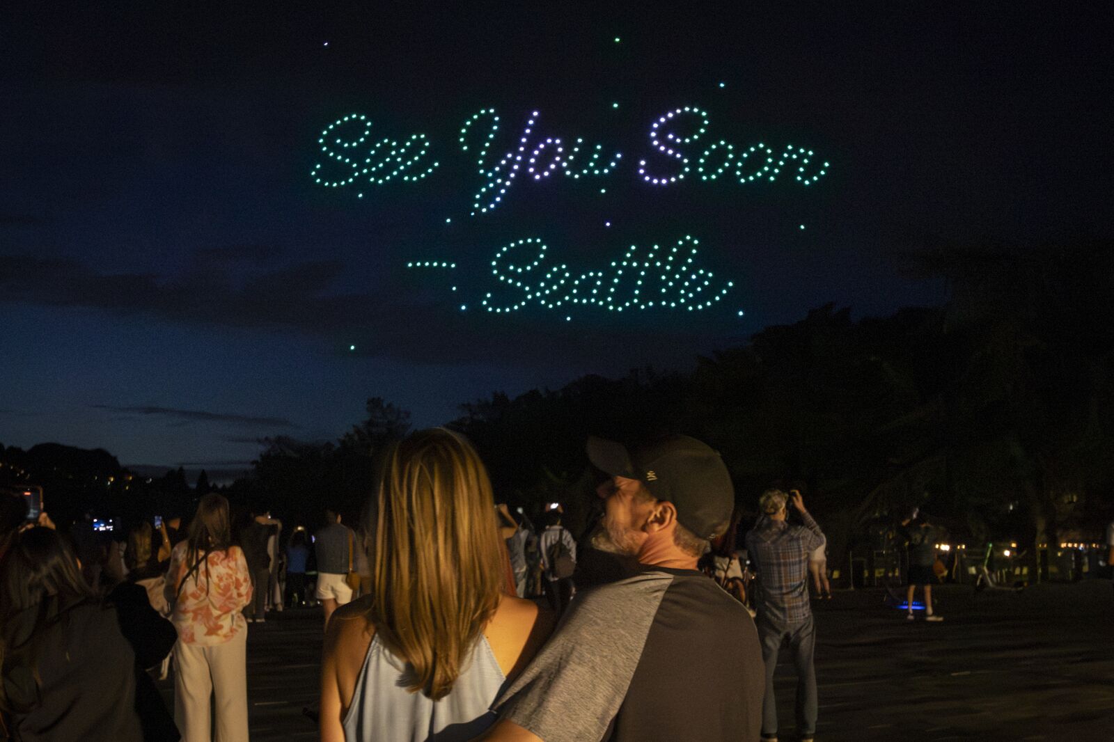 Photo prise au ras du sol au Seattle Center le soir d'un des matches de la Coupe du monde FIFA 26, groupe de spectateurs de dos et en silhouette qui regardent le ciel bleu nuit, au centre du cadrage un couple en léger contre-jour qui se serre, dans le ciel une inscription cursive lumineuse dessinée par des drones verts et violets formant les mots See You Soon Seattle en écriture manuscrite scintillante, un cliché intimiste bien différent du plan large sur la Space Needle