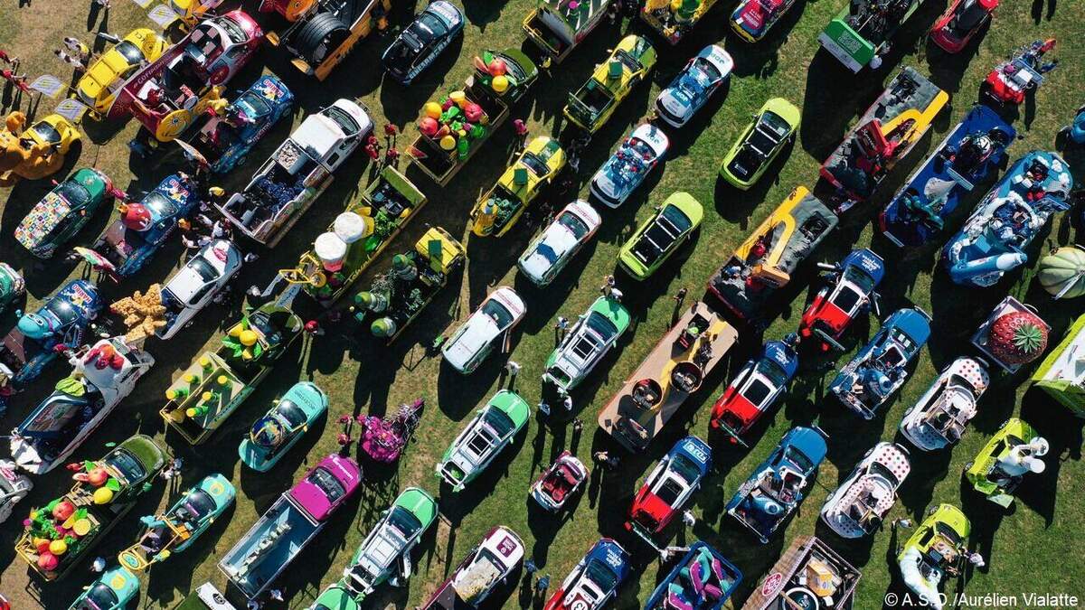 Photo aérienne prise en drone au-dessus d'un vaste champ vert où sont stationnés en rangs serrés plusieurs dizaines de véhicules publicitaires de la caravane du Tour de France 2026, camions et voitures aux formes détournées géantes empilant des mascottes colorées poulets rouges gâteaux jaunes pastèques bidons vert pomme et jaune canari boîtes carrées et parasols, allées régulières entre les cortèges avec silhouettes minuscules d'ouvriers en gilet jaune et casquettes, herbe verte lumineuse en fond, image documentaire prise avant le départ de Barcelone qui donne la mesure logistique du cortège