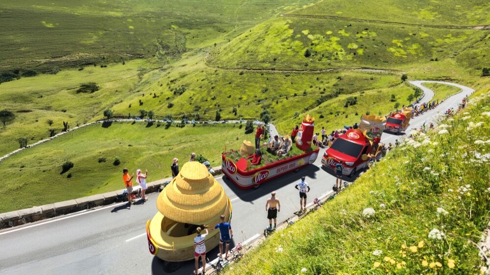 Photo prise sur route en pleine ascension pyrénéenne d'une étape du Tour de France 2026, cortège Vico en descente sur une route sinueuse au milieu de collines vertes flamboyantes et de bord de route herbeux fleuri, en tête un char sombrero jaune paille géant à roulettes vagues rouges tricolores latérales chapeau bombé mexicain, suivi d'un char plateforme rouge orné du logo Vico blanc et d'un sachet géant de chips Extra dorés, puis d'autres véhicules rouges alignés en file indienne, spectateurs debout au bord de la route en tee-shirt orange et blanc jambes nues, ciel bleu clair et bruyère éclairée par le soleil, scène en pleine étape opposée au plan large aérien de la cover