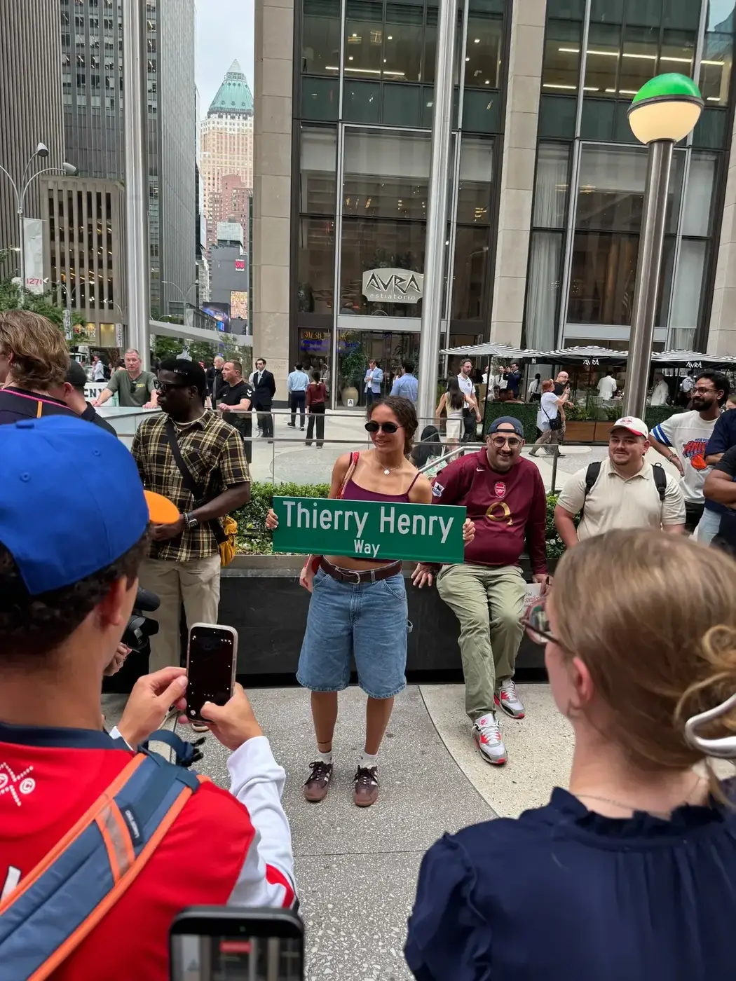 Photographie de la cérémonie d'inauguration de la Thierry Henry Way le 12 juin 2026 à Manhattan, la conseillère municipale Virginia Maloney présente le nouveau panneau bleu et blanc devant un parterre de supporters et de journalistes, façade en briques rouges typique du Midtown à l'arrière-plan, cérémonie officielle de la ville de New York