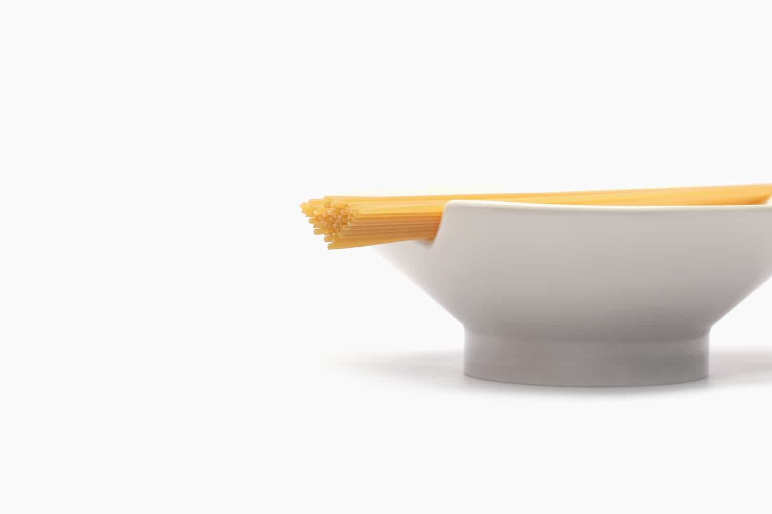 Photo studio en léger contre-plongée sur fond blanc lumineux de l'assiette à pâtes 80 Grammi signée Michele Venisti dévoilée le 16 juin 2026, coupe latérale sur une assiette creuse en porcelaine blanc mat au piédouche circulaire discret, sur son rebord supérieur droit un paquet vertical de spaghettis crus dorés glissé dans une encoche en V taillée dans le pourtour de l'assiette, la fibre de blé claire fait ressortir le glacis nacré de la céramique, image épurée qui met en scène la mesure comme un geste calme