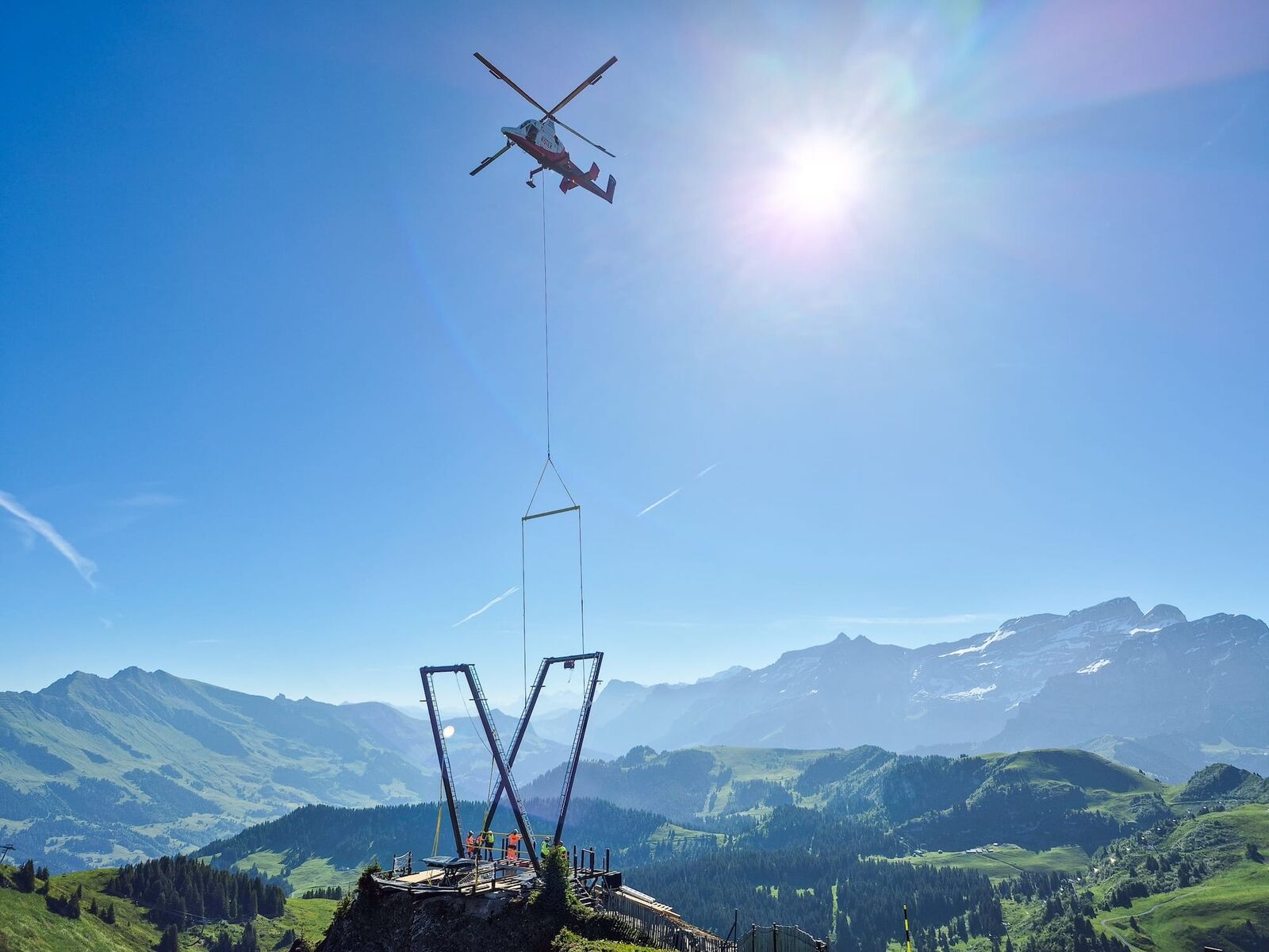 Photographie en plan large diurne prise sur un promontoire rocheux du Grand Chamossaire pendant le chantier de montage de la balançoire géante Balanc'AIR au printemps 2026, hélicoptère de transport de charges Kaman K-Max blanc et rouge de la compagnie suisse Rotex en vol stationnaire au centre du cadre suspendant une longue élingue verte à laquelle est attaché un pylône métallique noir en cours de dépose sur une plateforme en construction en bois clair au sommet de la crête, deux ouvriers en gilet orange fluo attendent la charge en contrebas, panorama grandiose sur les Alpes vaudoises et suisses avec sommets pointus enneigés du massif des Diablerets à droite pâturages verts en contrebas et vallée du Chablais dans la brume matinale, ciel bleu vif avec flare solaire éclatant en haut à droite