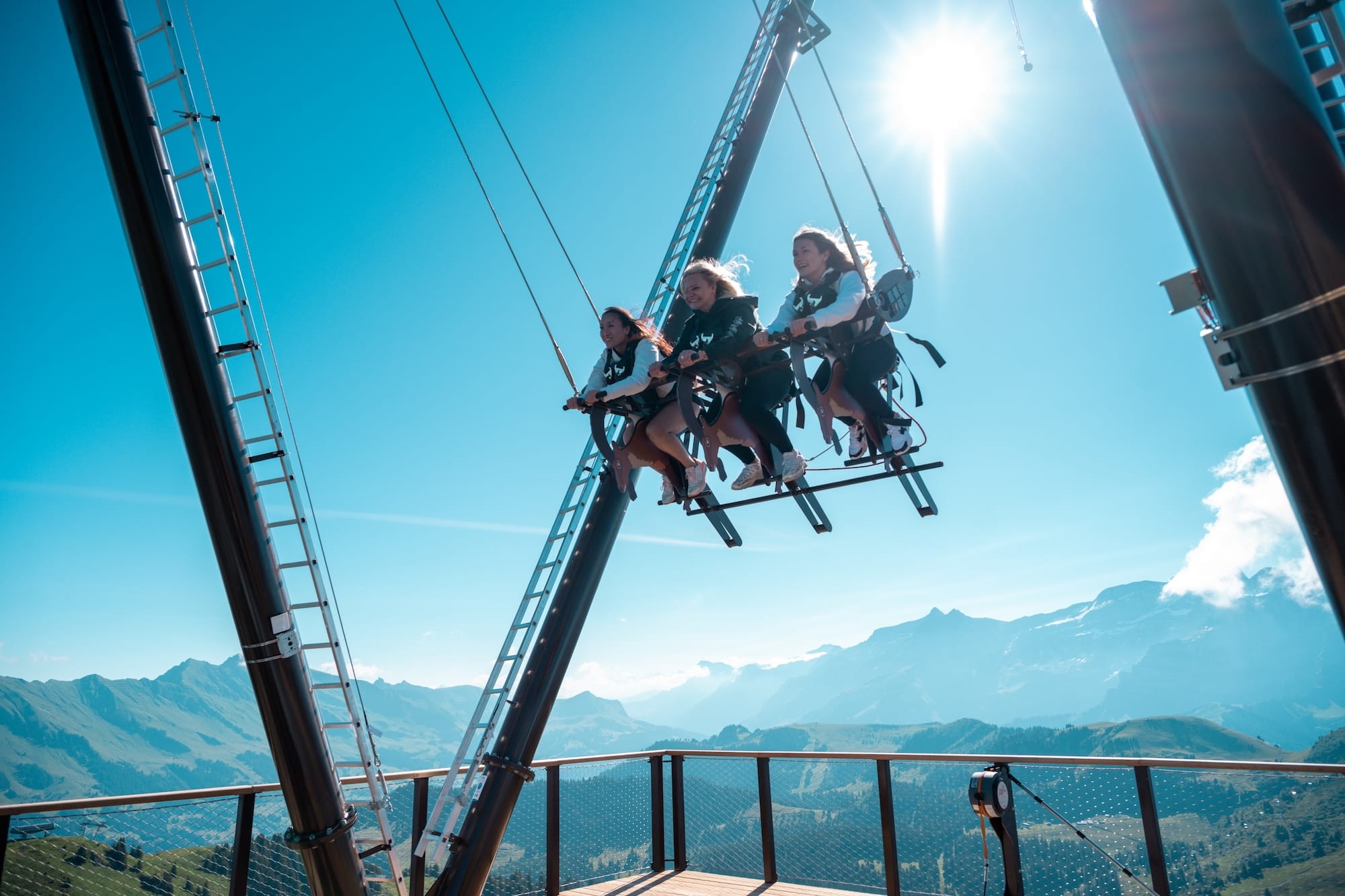Photographie diptyque diurne de l'attraction Balanc'AIR inaugurée le 3 juillet 2026 à Villars-Gryon-Diablerets dans les Alpes vaudoises, à gauche plan large plein soleil sur la structure métallique noire en V renversé haute d'une dizaine de mètres plantée sur une plate-forme en bois clair au sommet de la crête herbue du Grand Chamossaire à 2110 mètres, trois personnes assises côte à côte harnachées sur la nacelle en pleine bascule dans le ciel bleu vif face à la silhouette découpée du sommet des Dents-du-Midi enneigées, à droite plan serré en contre-plongée de trois jeunes femmes cheveux au vent en sweat noir sur la nacelle éclatant de rire un halo solaire baignant la scène, cadrage horizontal image officielle de la station