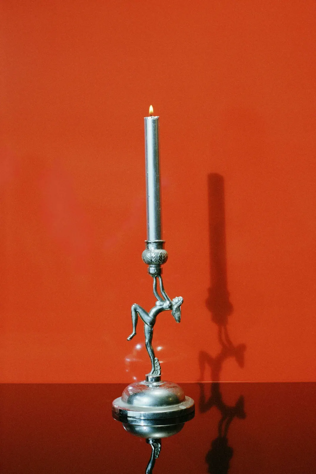 Photographie Candleholder woman de Camille Vivier datée de 2021 et présentée dans sa rétrospective MEP du 10 juin 2026, nature morte sur fond rouge orangé saturé montrant un bougeoir en argent ouvragé sculpté en forme de femme nue dansante portant une bougie blanche allumée sur sa tête, projection d'ombre théâtrale sur le mur