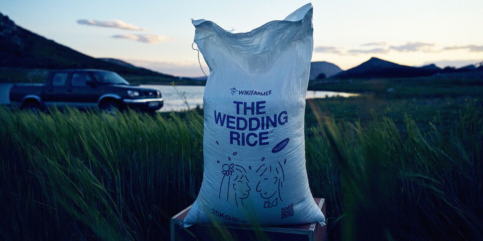 Photo de production réalisée par le photographe Panos Pagonis pour la campagne The Wedding Rice de Wikifarmer dévoilée par McCann Worldgroup Athènes le 11 avril 2025, gros sac de riz blanc de 25 kg posé sur une caisse en bois au milieu d'un champ d'orge encore vert tendre, pickup gris stationné au loin sur un chemin de terre devant les eaux d'un lac, montagnes basses se découpant sur un ciel bleu profond de fin de journée