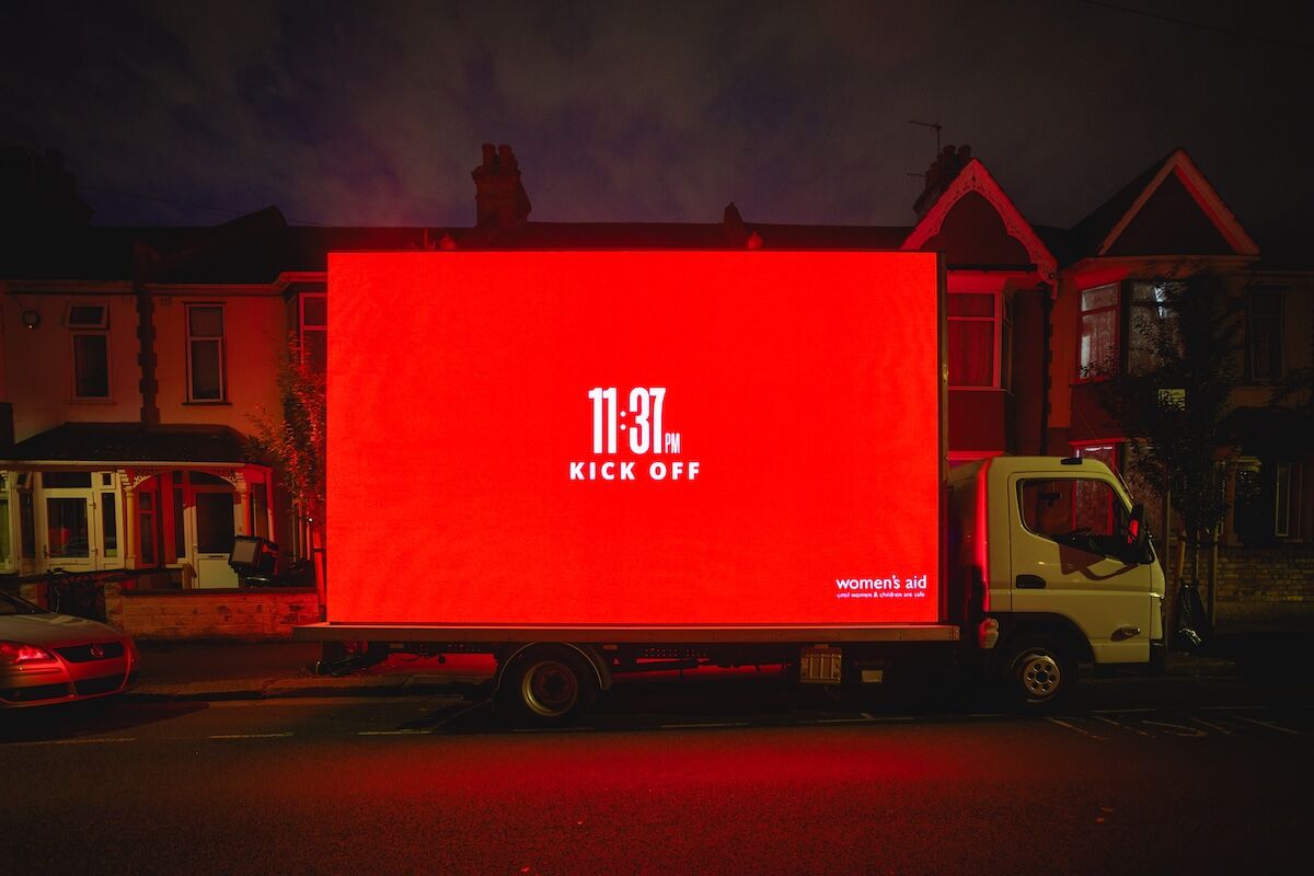 Photo de nuit prise dans une rue résidentielle du nord de Londres pendant la campagne The Other Kick Off signée Elvis pour Women's Aid le 18 juin 2026, mobile billboard sur un camion à plateau garé le long de maisons victoriennes en brique rouge et tuile ardoise, immense écran rouge saturé rétroéclairé affichant en typographie sans serif blanche l'inscription 11 37 PM Kick Off et le logotype women's aid en bas à droite avec la mention until women and children are safe, une voiture rouge en premier plan, ciel gris pommelé, ambiance d'alerte silencieuse qui vire le rouge criard des stades en signal de détresse domestique