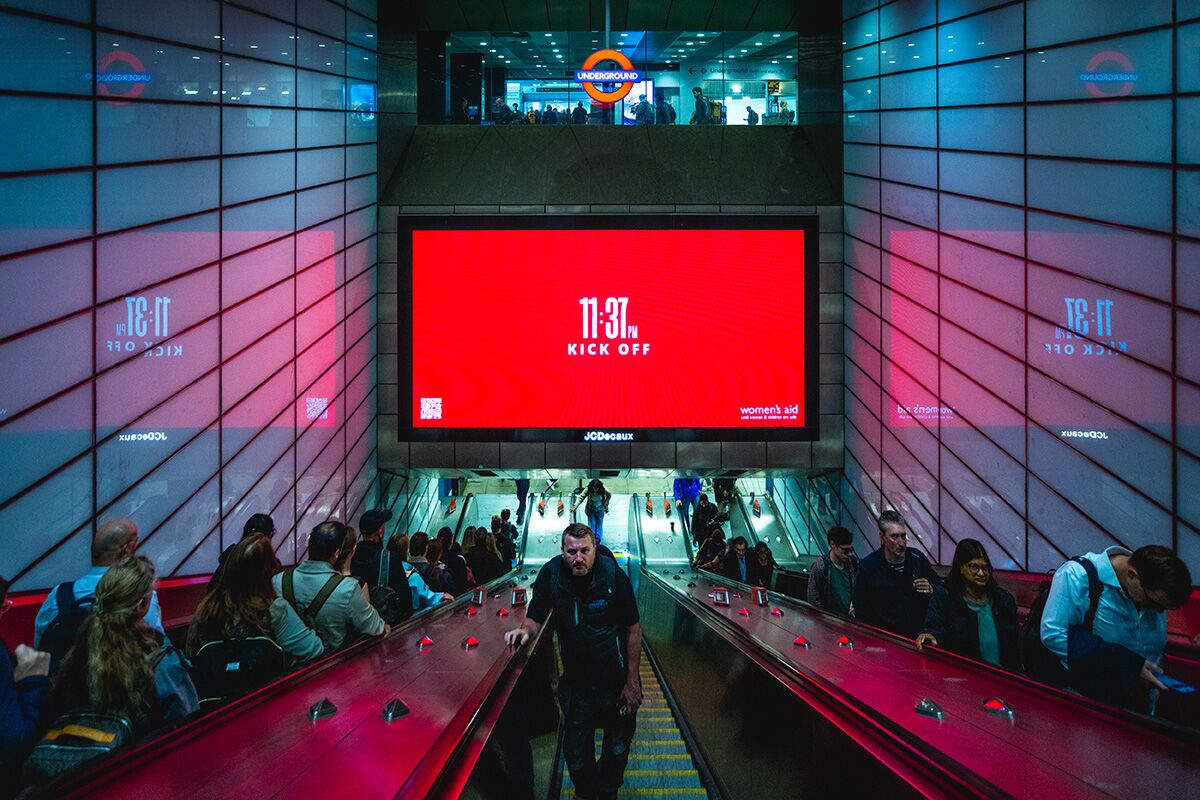 Photo prise dans une station du métro londonien pendant la campagne The Other Kick Off de Women's Aid, cadrage frontal sur les escalators rouges d'un accès à la ligne Underground, panneau digital JCDecaux central écran rouge saturé affichant 11 37 PM Kick Off en typographie blanche et logo women's aid, reflets rétroéclairés du visuel qui se répètent sur les parois latérales du métro en surimpression fantomatique de l'inscription 11 37 KICK OFF, une trentaine de voyageurs qui remontent et descendent les escalators dans une lumière crue, écriture Underground orange visible en haut