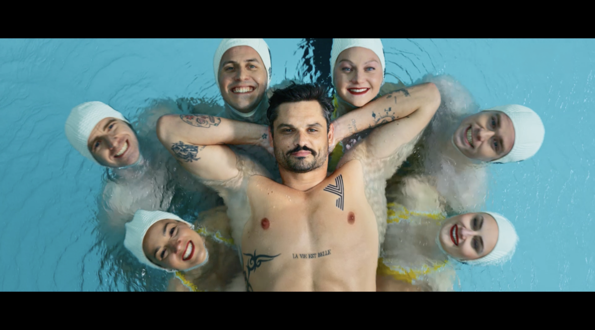Photogramme du film Le Ballet de Yomoni signé Brainsonic et réalisé par ARGO, plan aérien vertical en plongée sur la surface d'un bassin de la piscine Molitor à Paris, Florent Manaudou allongé au centre sur le dos les yeux fermés torse nu à peau claire mains croisées derrière la nuque tatouages tribaux et inscription La vie est belle sur le buste, six membres de l'équipe de France de nage synchronisée disposés autour de lui en couronne symétrique tête en avant bonnets de bain blancs souriants deux à droite deux à gauche et deux en haut de la formation, eau bleu turquoise miroitante avec reflets clairs, cadrage cinématographique bord noir en haut et en bas format cinéma