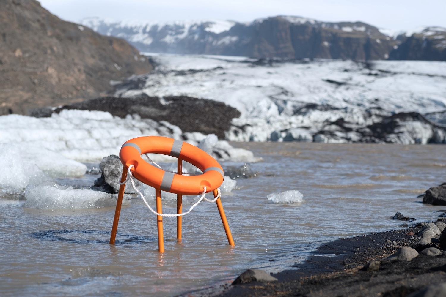 Photographie diurne en plan large paysage de la Sea Level Rise Chair du designer italien Tobia Zambotti installée en Islande début juillet 2026, chaise upcyclée à structure métallique fine peinte en orange vif à quatre pieds inclinés dont deux pieds arrière plongent dans l'eau glacée boueuse d'un ruisseau glaciaire, assise composée d'une bouée de sauvetage circulaire orange en polyéthylène marquée de deux bandes réfléchissantes grises et d'une corde blanche tressée qui pend, arrière-plan spectaculaire d'une langue de glacier grise et blanche craquelée entrelacée de striures de cendres volcaniques noires, montagnes rocheuses claires en fond dans une brume légère, sol de galets sombres à droite du cadre, lumière naturelle rasante et diffuse d'un ciel voilé